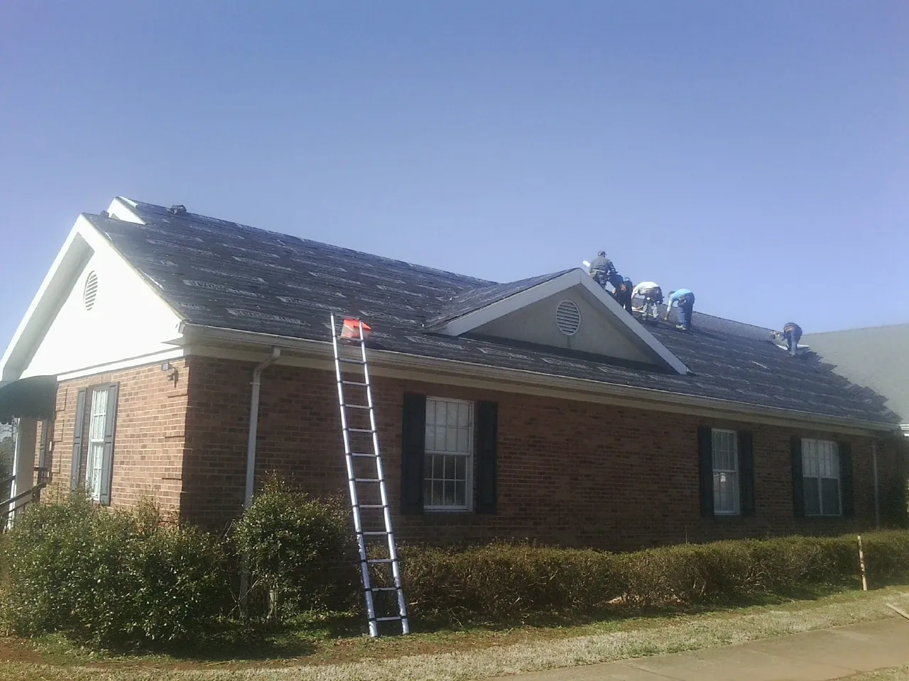 Crew tearing shingles off a brick ranch roof with a ladder set — Metal Roof Repair work by our crew in Olsburg