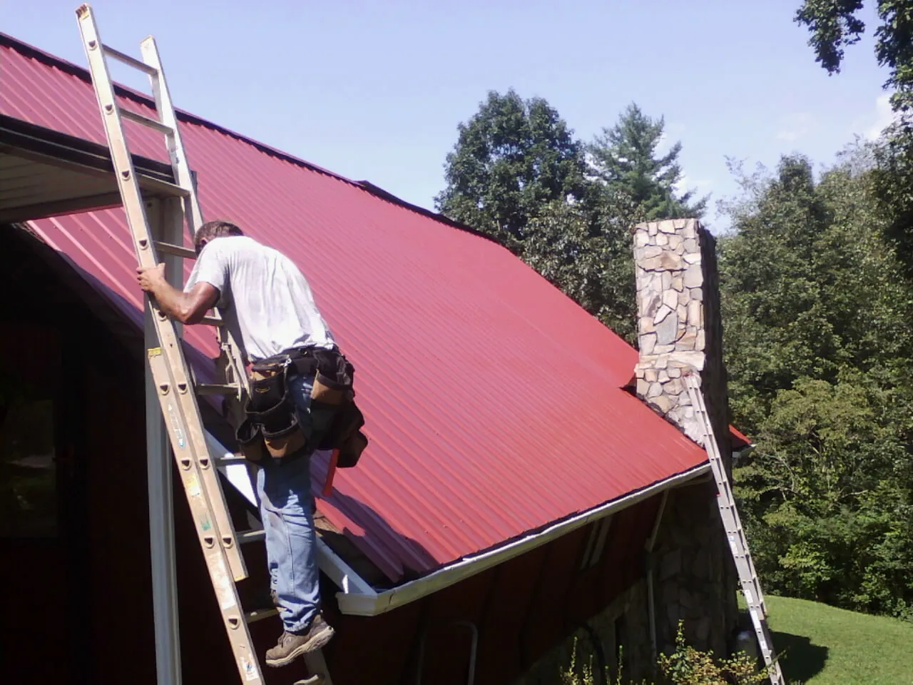 Roofer on a ladder beside a red metal roof and stone chimney — Soffit & Fascia Repair by Roofing Brothers in Olsburg
